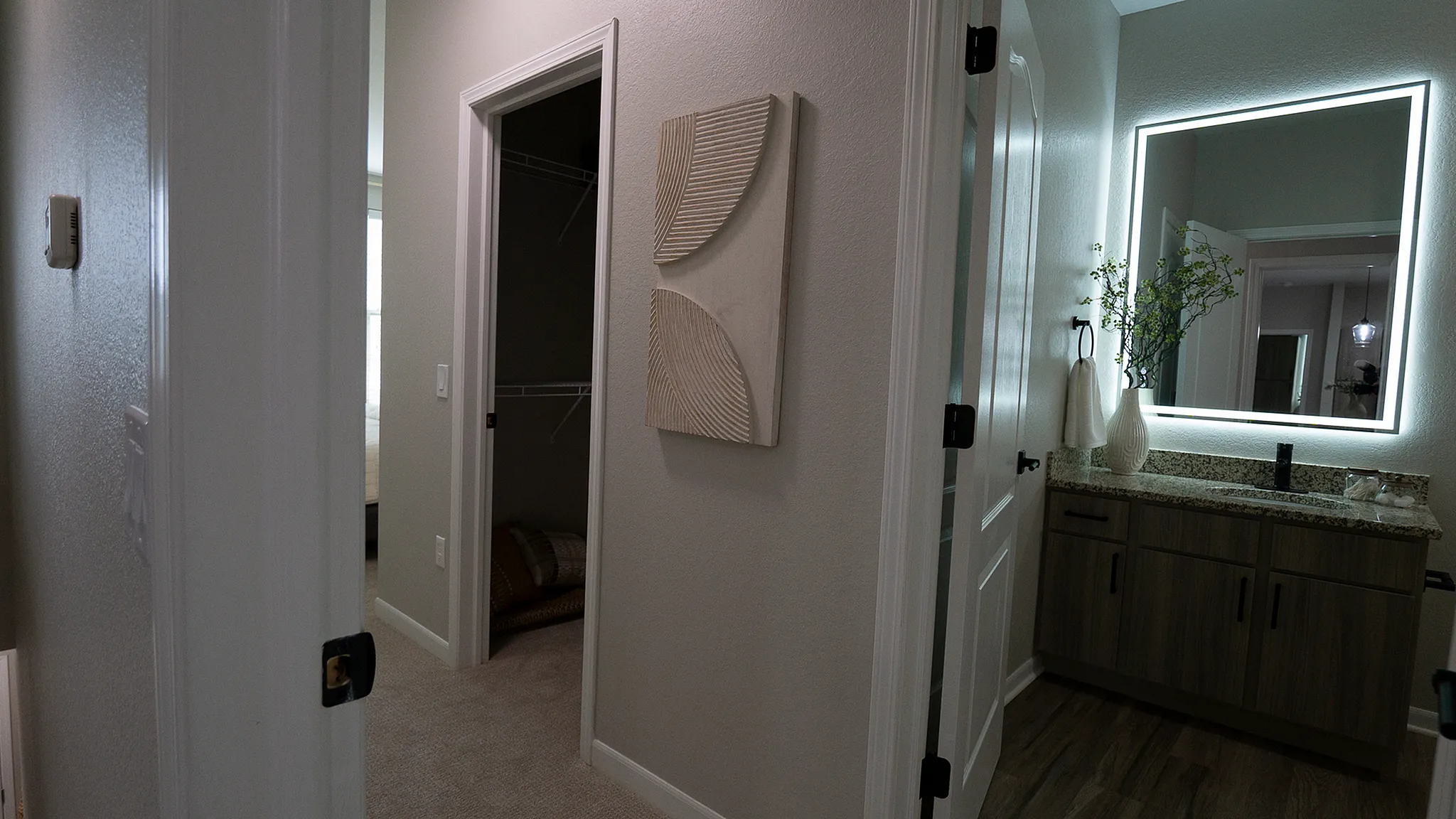 Apartment hallways connecting a spacious bedroom closet and renovated bathroom with large, illuminated mirror.
