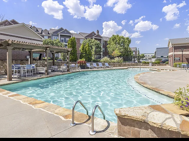 Inviting outdoor pool area with crystal-clear water, sun loungers, shaded seating, and vibrant landscaping against a backdrop of stylish apartment buildings.