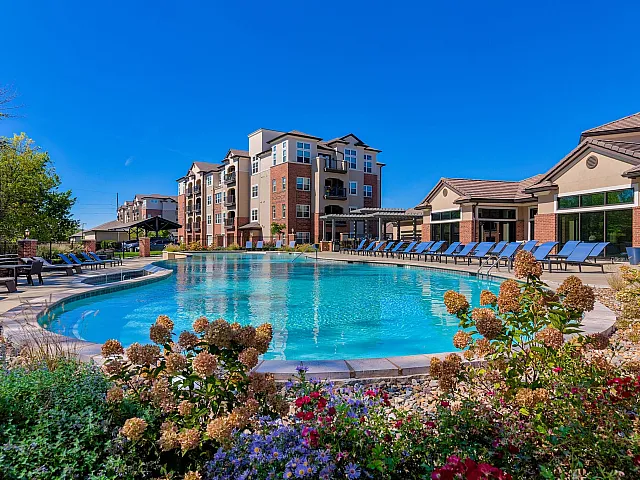 Luxurious apartment complex featuring a resort-style pool surrounded by lush landscaping and comfortable lounge chairs under a clear blue sky.