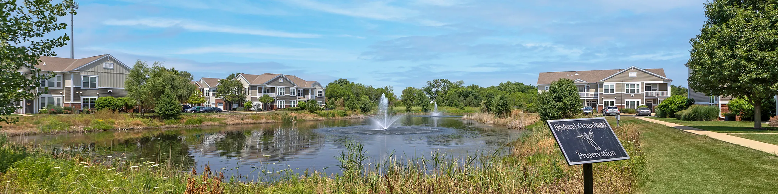 Charming apartment community with serene pond view, fountains, and lush greenery in a natural grassland setting.
