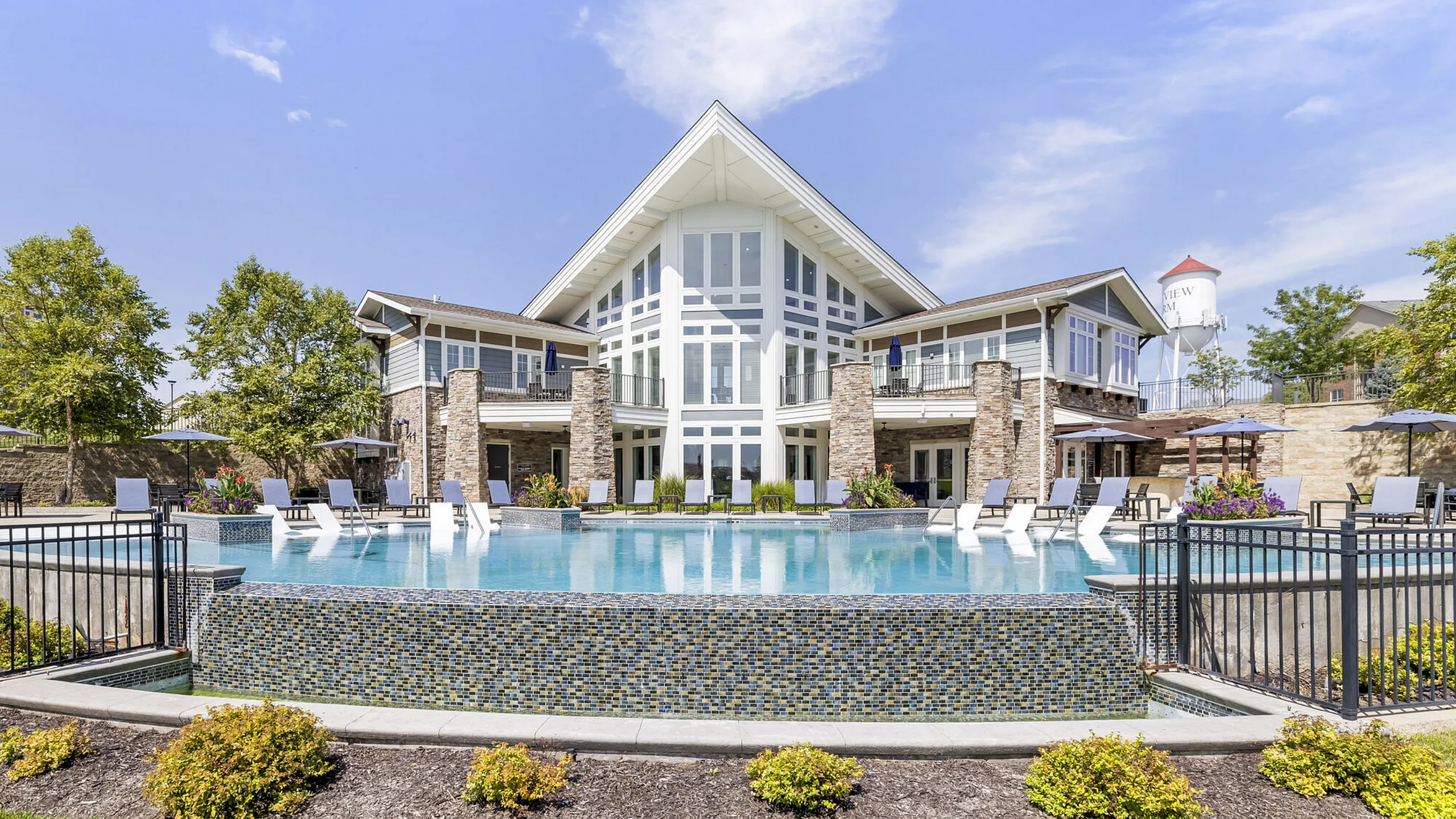 Luxurious apartment complex featuring a stunning pool with lounge chairs, surrounded by elegant stone architecture and lush landscaping on a sunny day.