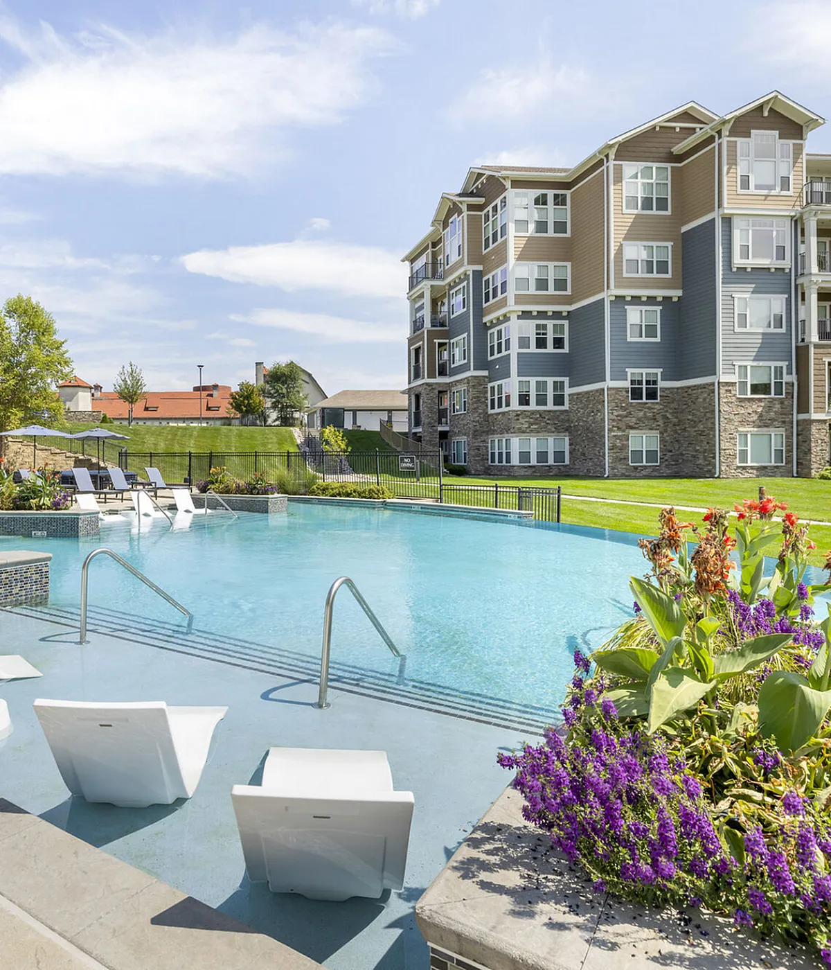 Modern apartment complex with a sparkling pool, surrounded by lush greenery and colorful flowers, offering a serene outdoor escape.