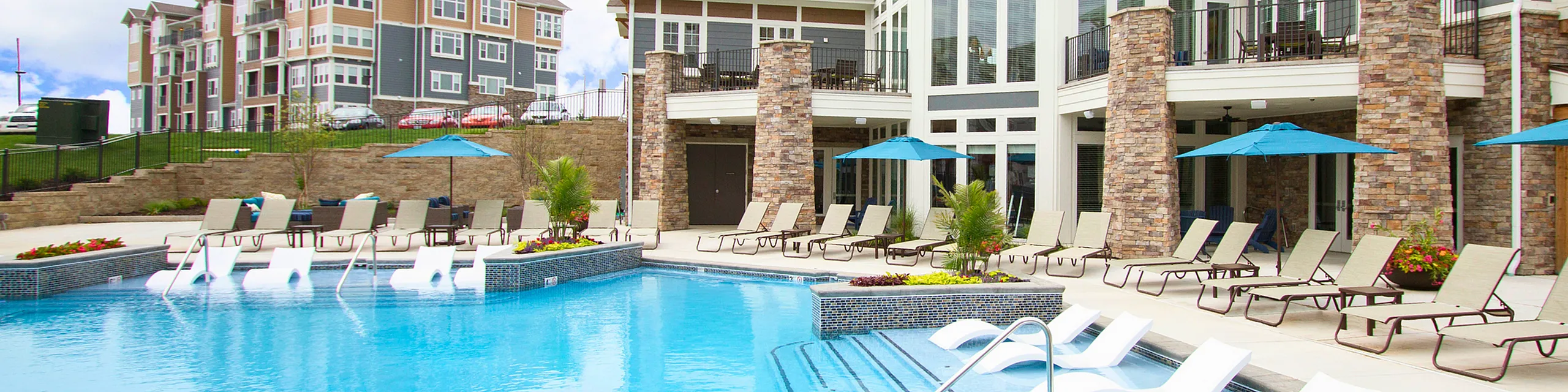 Luxurious apartment complex with a sparkling pool, stylish lounge chairs, and elegant stone architecture under bright blue skies.
