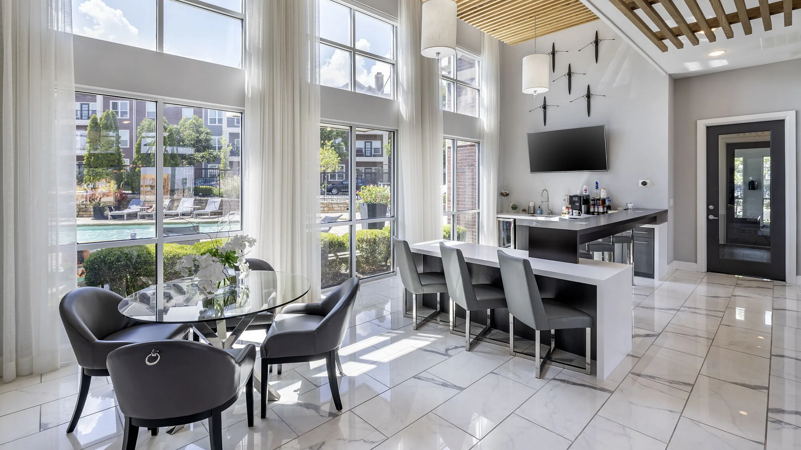 Elegant lounge with floor-to-ceiling windows, marble tiles, and a chic bar area, overlooking a sunlit poolside retreat.