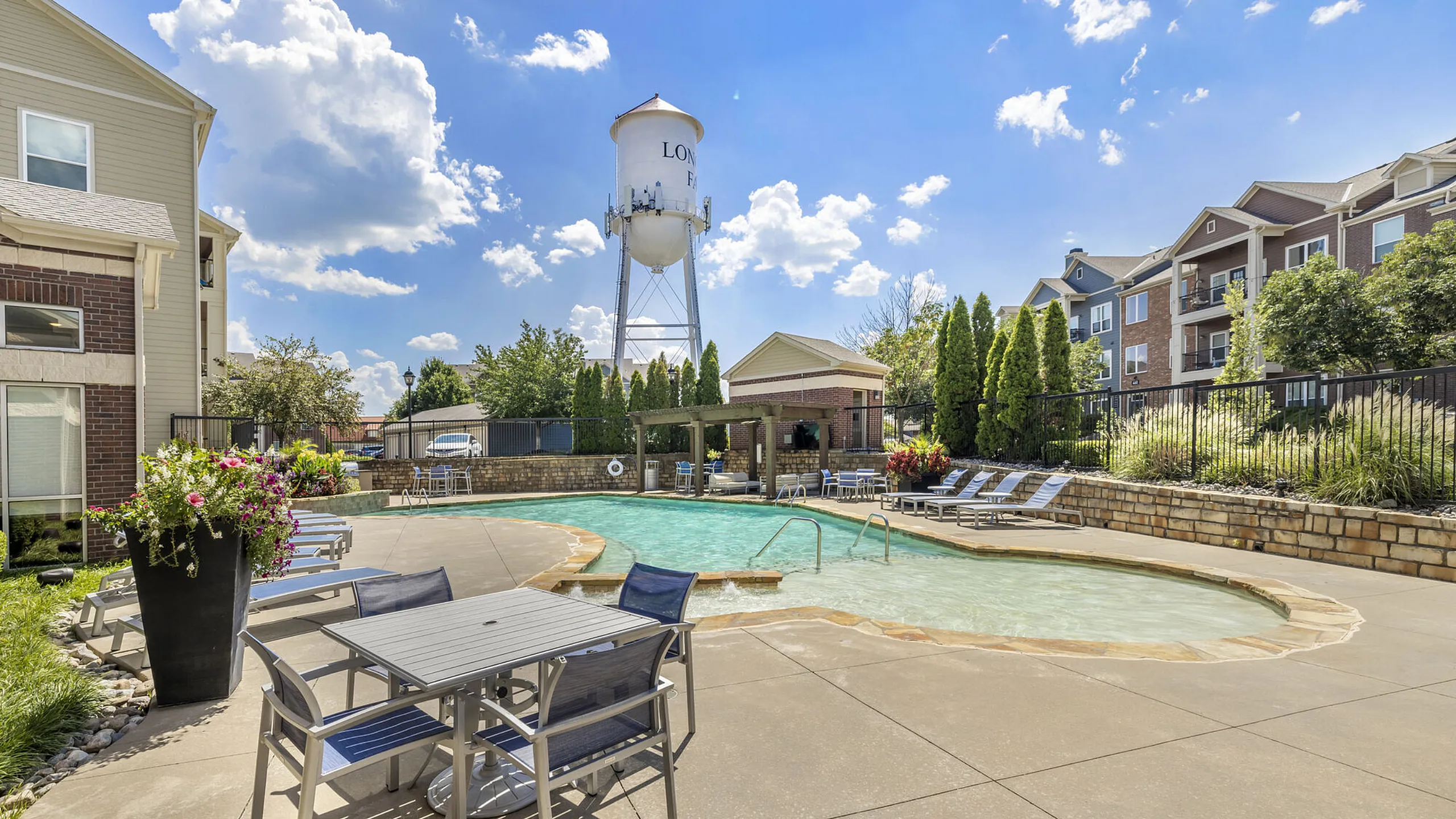 Charming apartment complex with a sparkling pool, surrounded by lush landscaping and modern outdoor seating, under a bright blue sky.