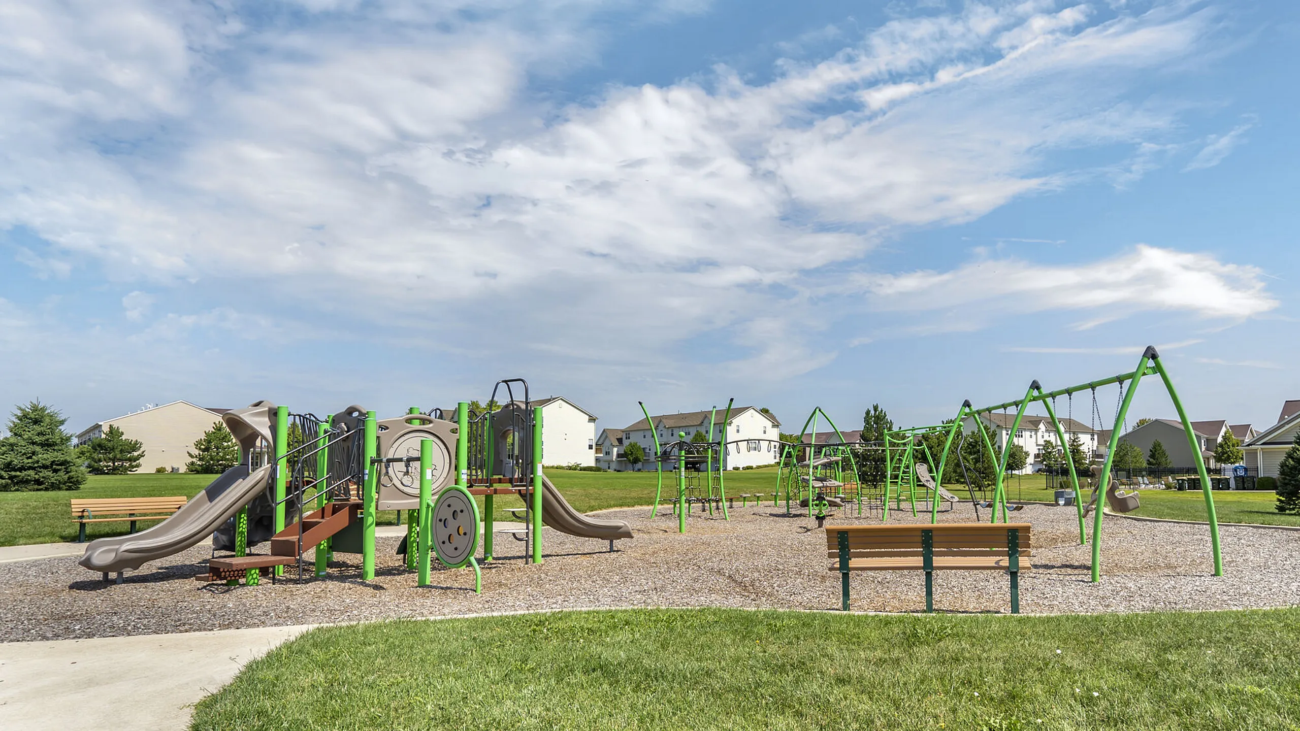 Vibrant community playground with modern swings and slides, surrounded by lush green grass and family-friendly homes in the background.
