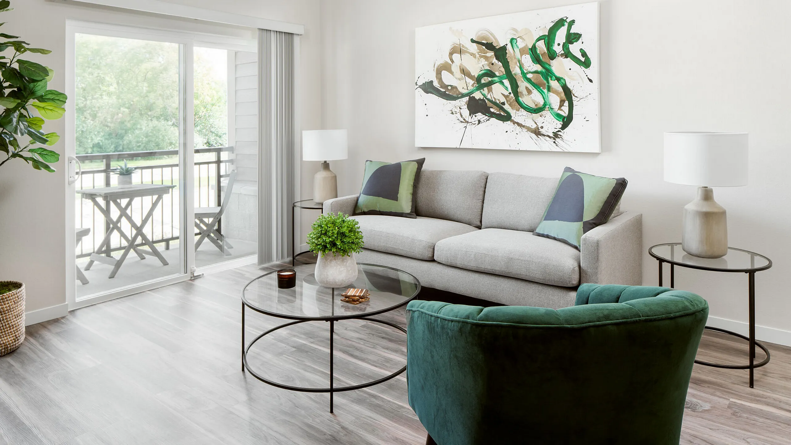 Modern living room with a cozy gray sofa, stylish glass coffee table, and abstract art, opening onto a bright balcony with lush greenery.