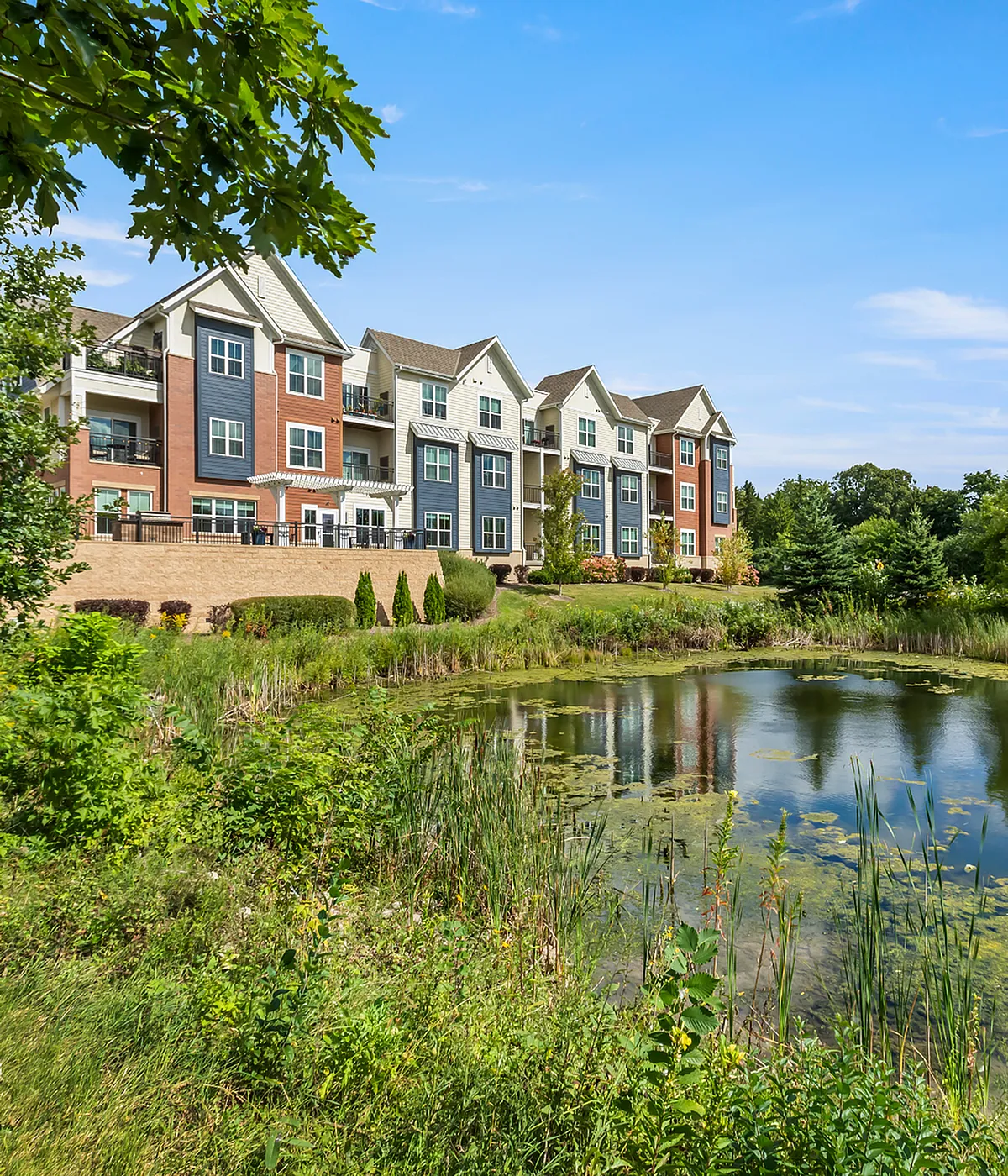 Charming apartment complex with modern architecture set amidst lush greenery and a serene pond view.