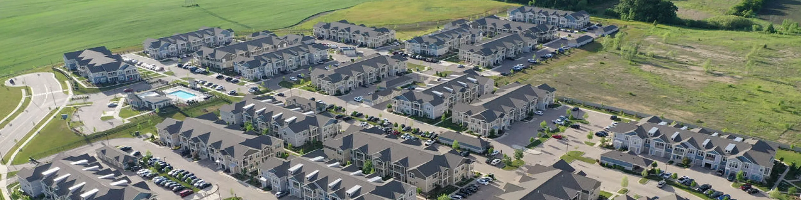 Charming aerial view of a modern residential complex with lush green surroundings, ample parking, and a refreshing community pool.
