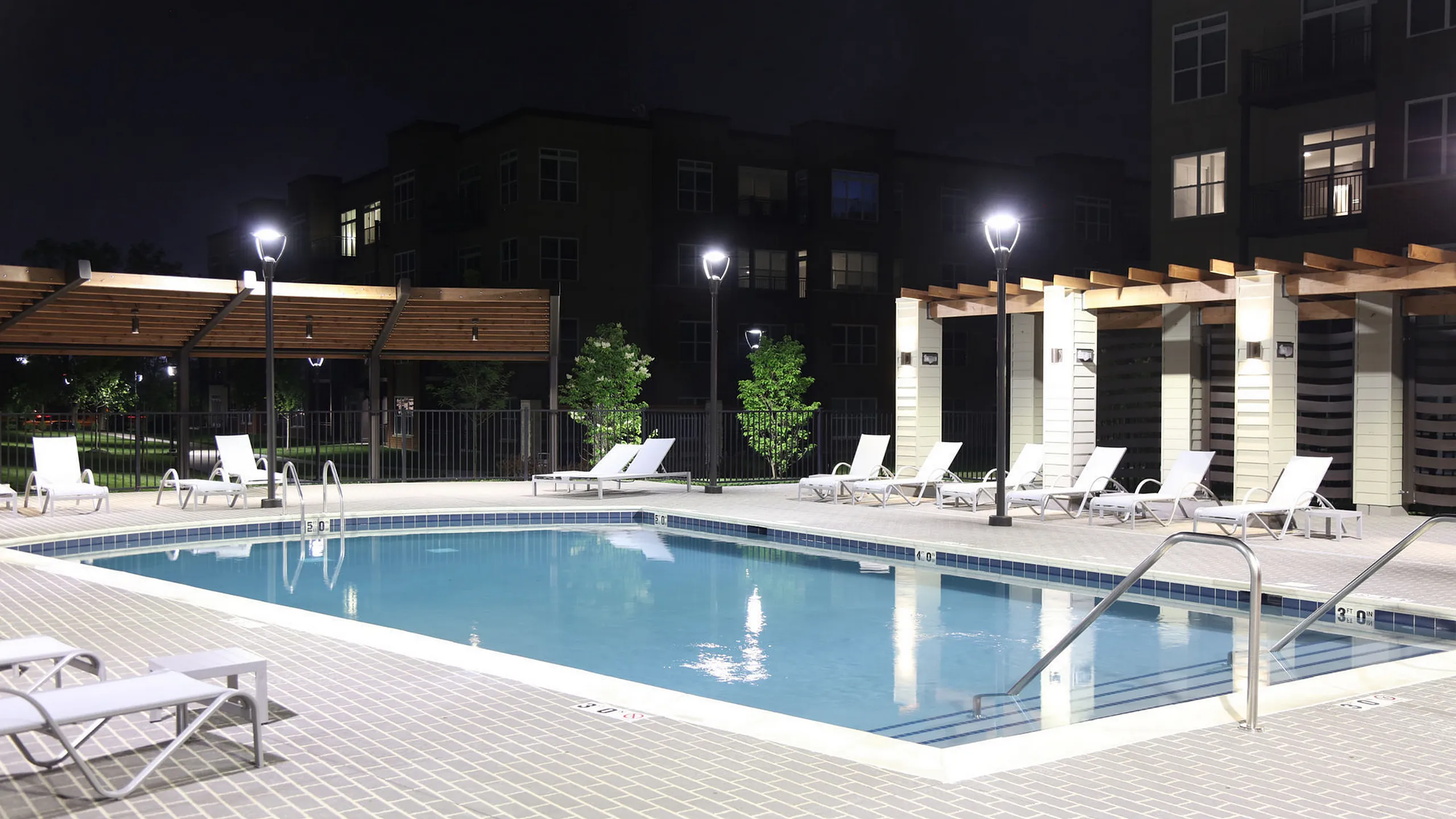 Luxurious outdoor swimming pool area with elegant lounge chairs and stylish pergolas, beautifully illuminated for nighttime relaxation.
