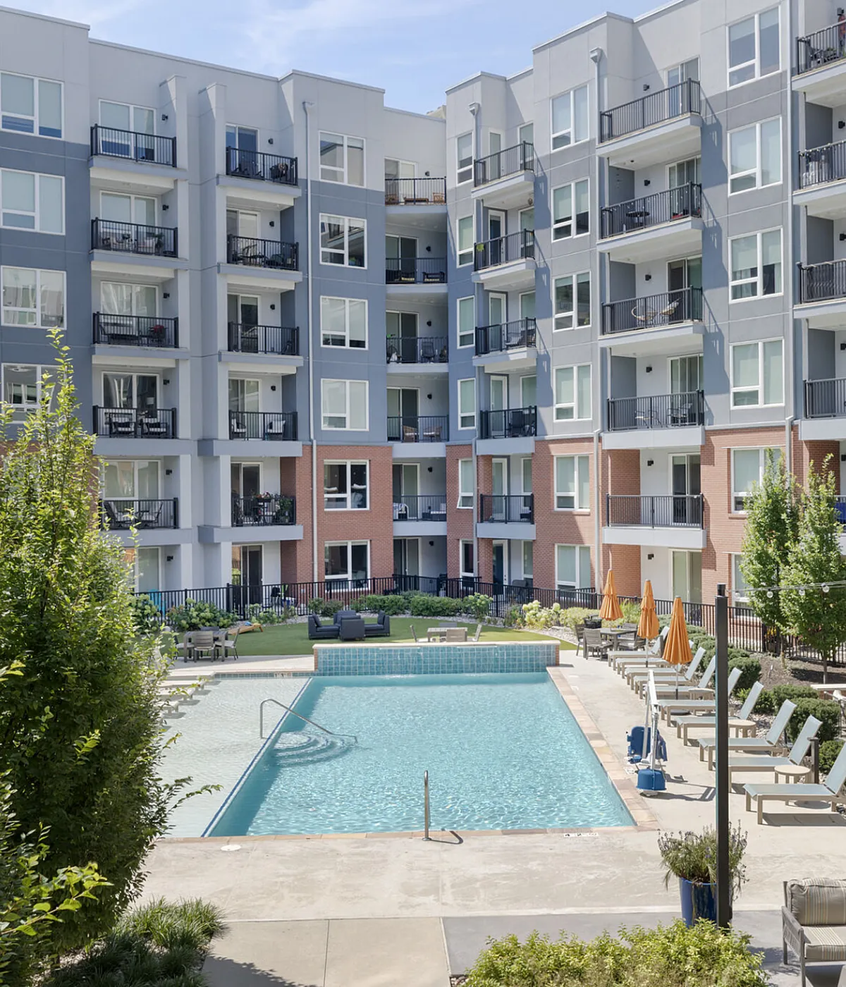 Elegant apartment complex with modern balconies overlooking a sparkling pool, surrounded by lush greenery and outdoor seating areas.