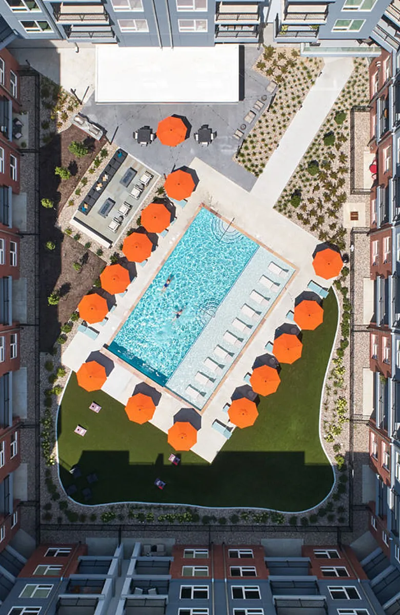 Aerial view of modern apartment complex featuring a refreshing outdoor pool with vibrant orange umbrellas, lush green landscaping, and stylish seating areas.