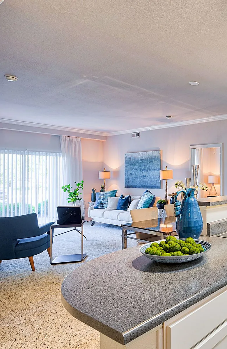 Bright and modern living area with chic decor, featuring a cozy white couch, elegant blue accents, and a lush view from large windows.