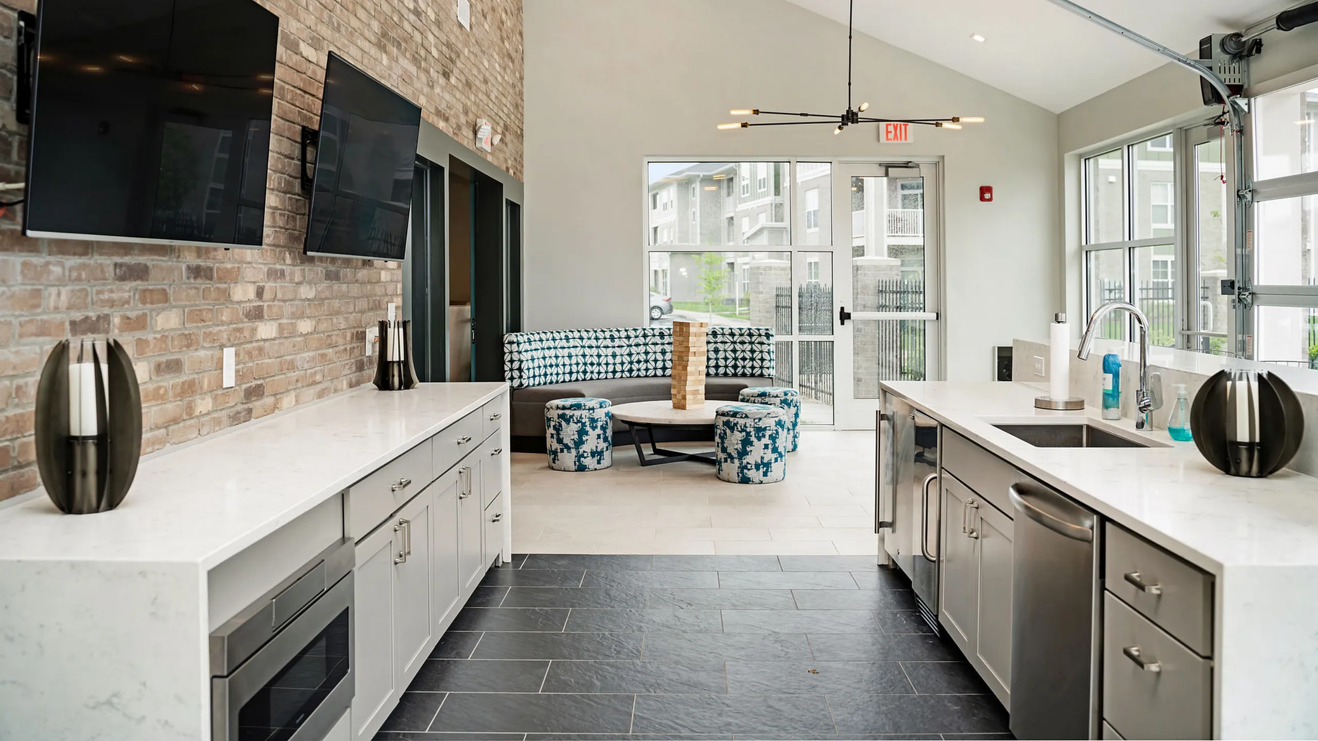 Modern communal kitchen area with sleek white countertops, dual sinks, stainless steel appliances, and a cozy seating nook featuring patterned furniture and a large window for natural light.