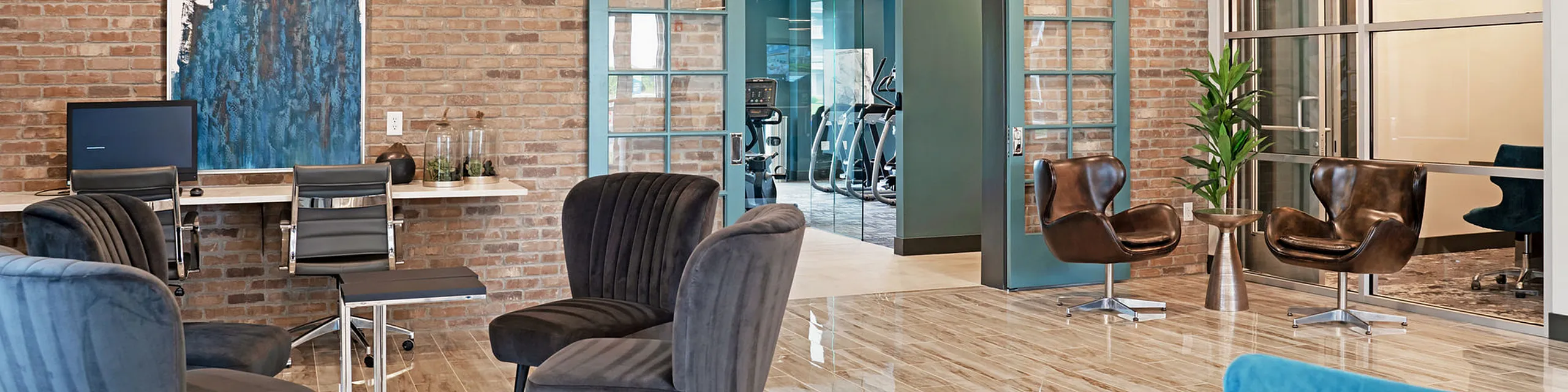 Modern communal lounge with stylish seating, exposed brick walls, and a view into a well-equipped fitness center.