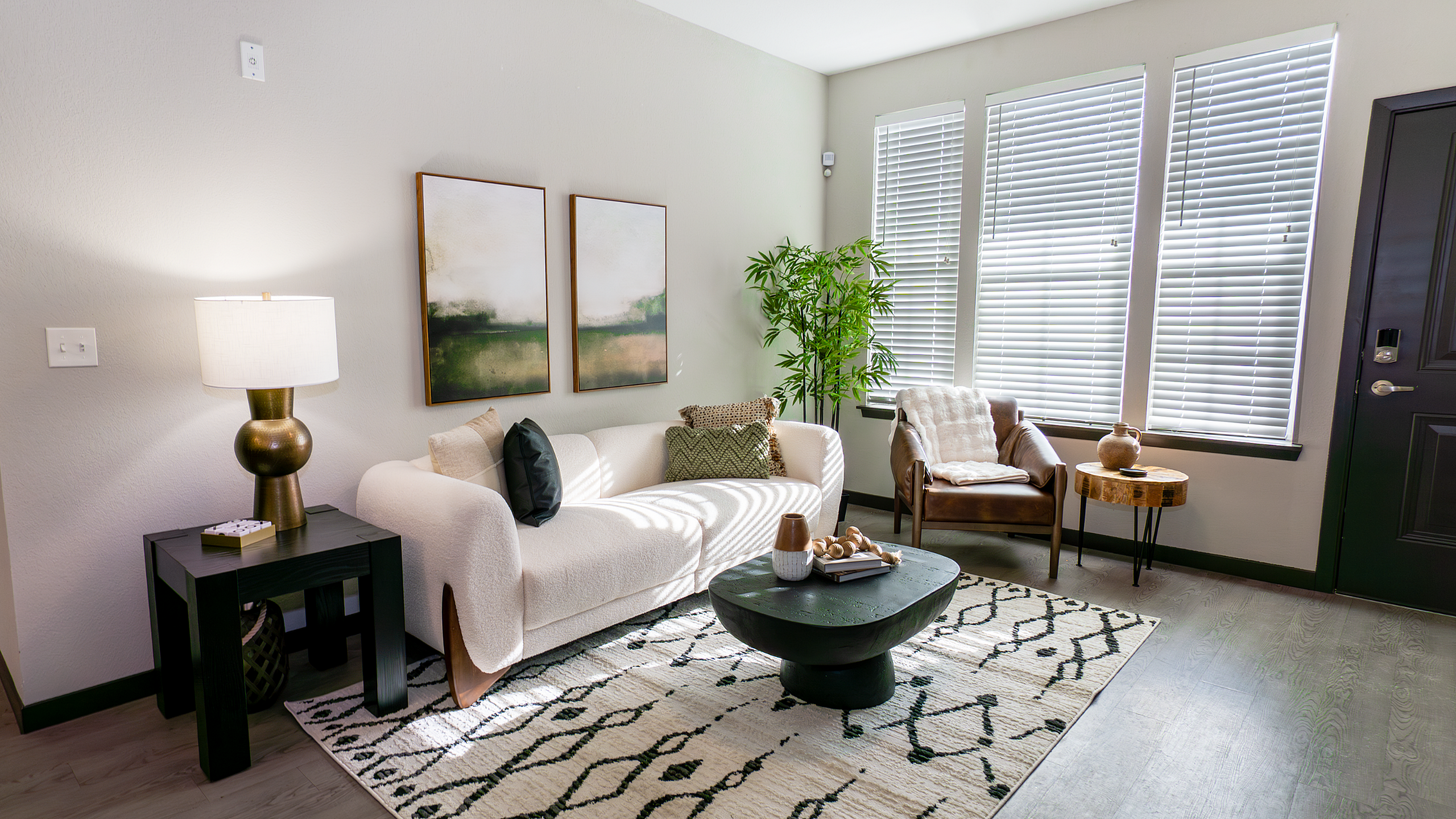 Bright and inviting living room with chic decor, featuring a plush white sofa, elegant black side table, and natural light streaming through large windows with blinds.