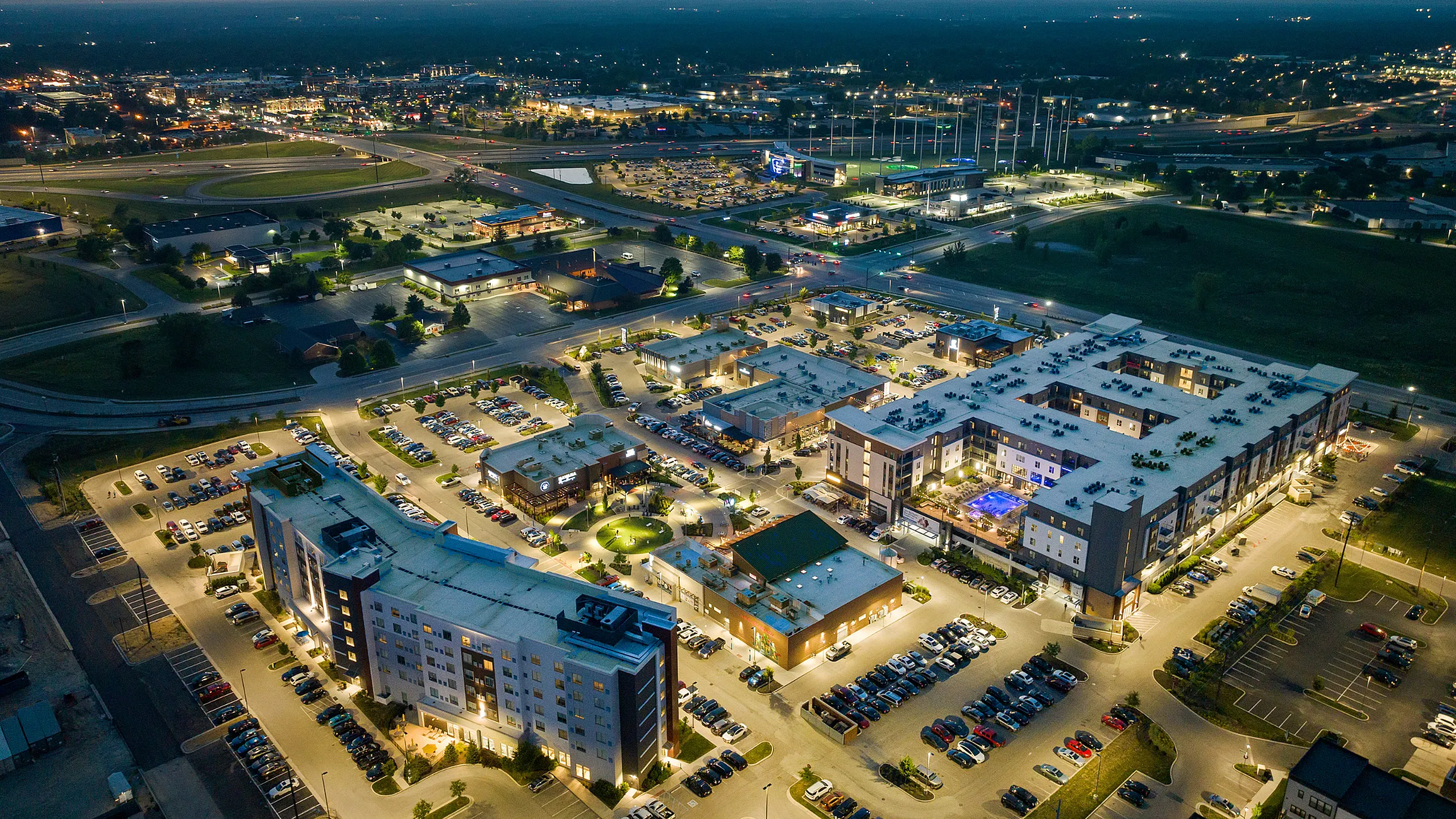 Aerial view of a vibrant, illuminated apartment complex featuring modern architecture, surrounded by ample parking and retail spaces in a bustling neighborhood.