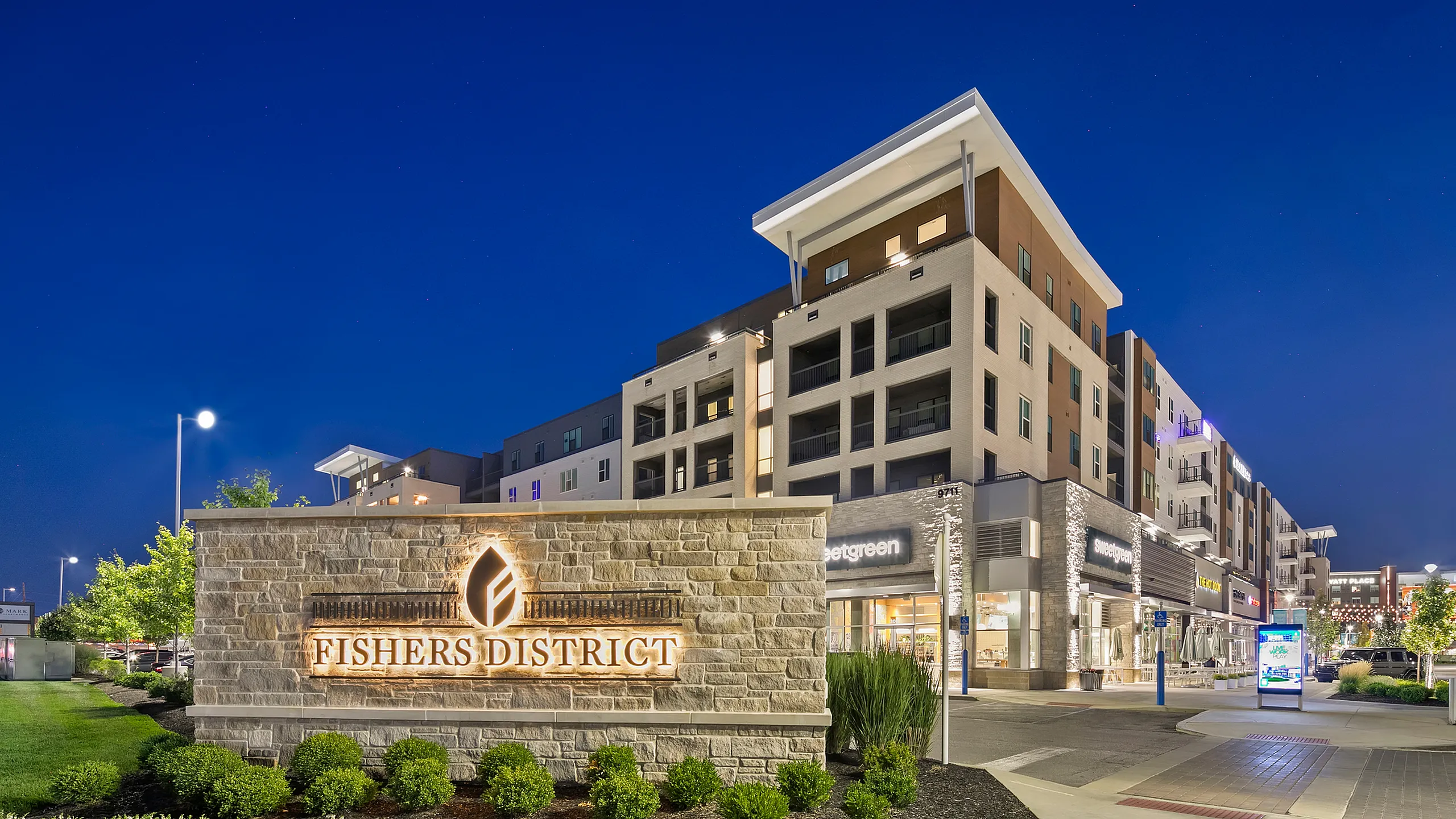 Modern apartment complex at Fishers District illuminated at night, featuring retail shops and lush landscaping.