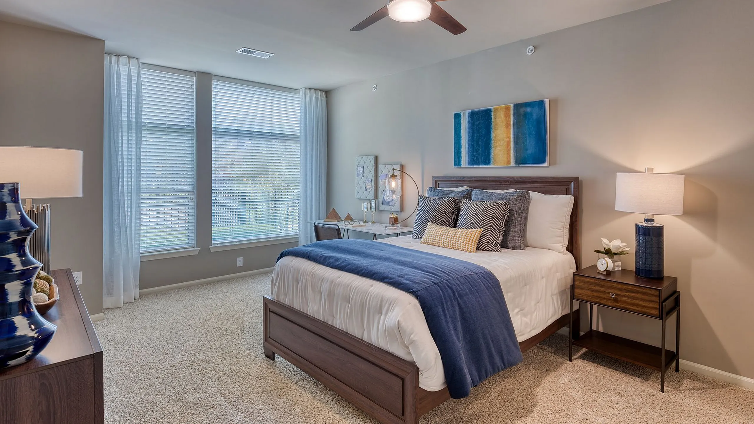 Elegant bedroom with modern decor, featuring a plush bed with blue accents, large windows with natural light, and stylish furniture for a cozy, inviting atmosphere.