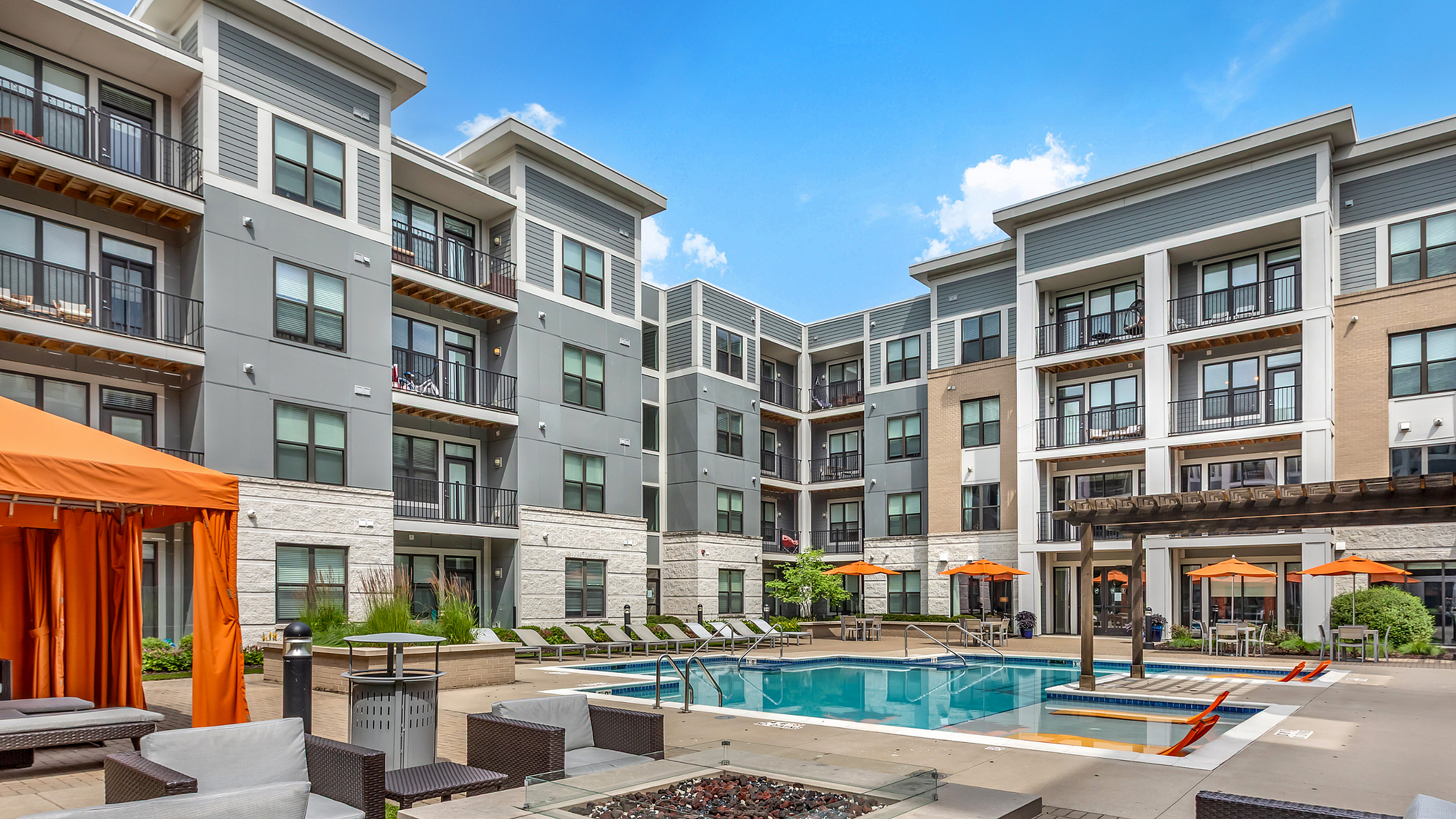 Modern apartment complex featuring sleek architecture with private balconies, vibrant pool area, and luxurious lounge seating under bright orange canopies.