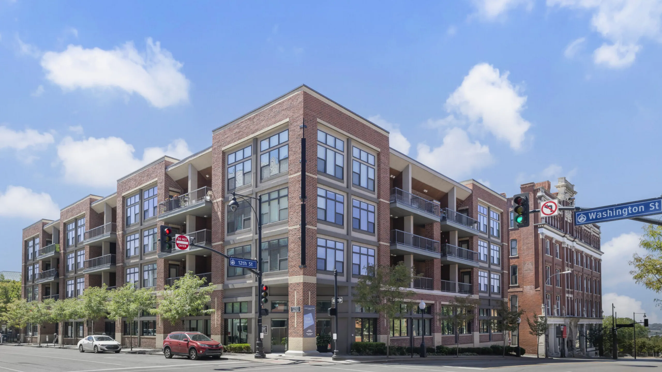 Modern apartment complex with brick facade and large windows, located on a sunny street corner, offering urban living with convenience and style.