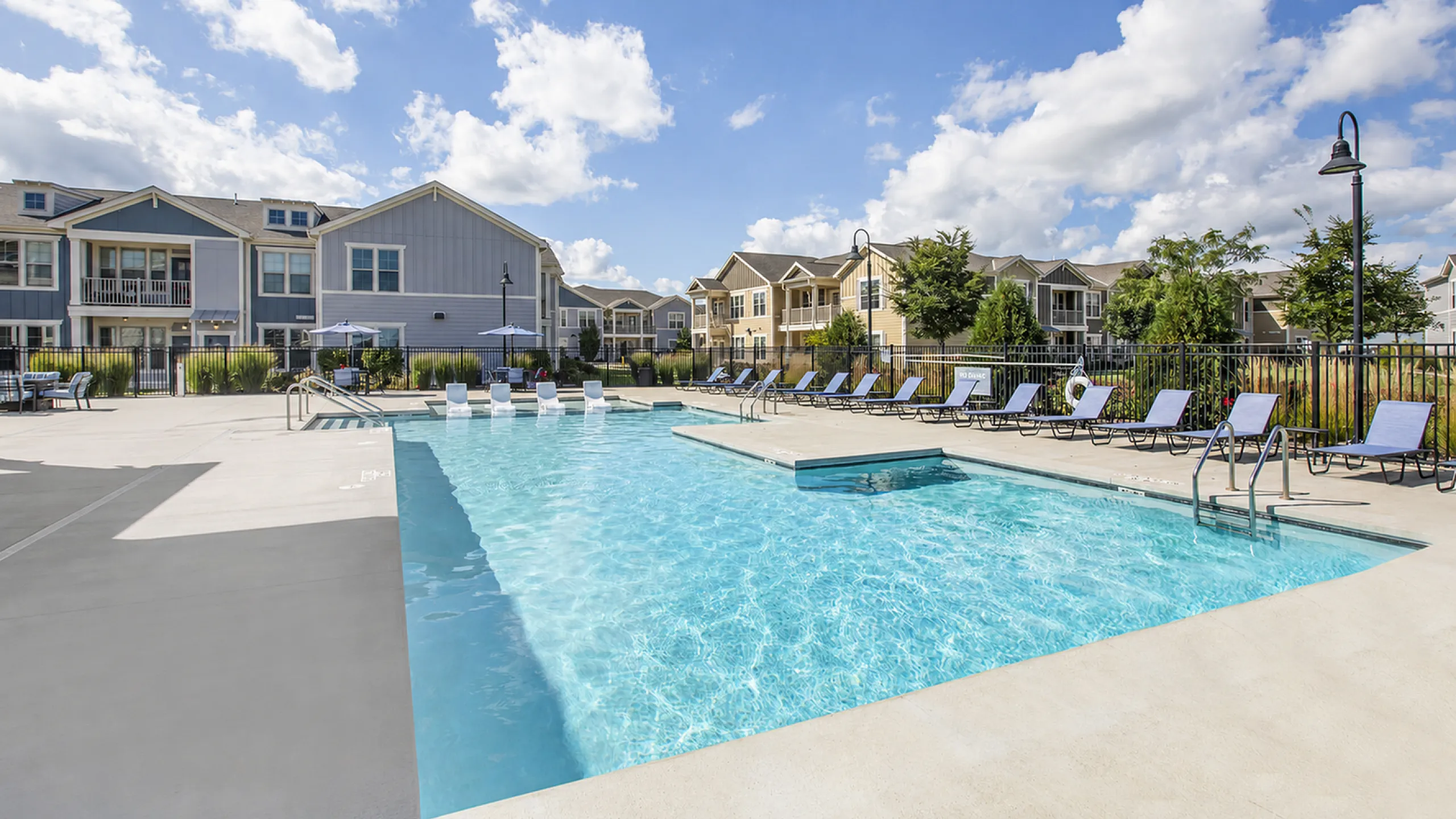 Inviting swimming pool area with comfortable lounge chairs, surrounded by stylish apartment buildings under a bright blue sky.