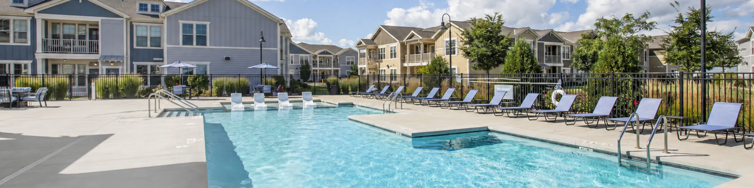 Inviting apartment complex with a sparkling swimming pool, ample lounge chairs, and charming architecture under a bright blue sky.