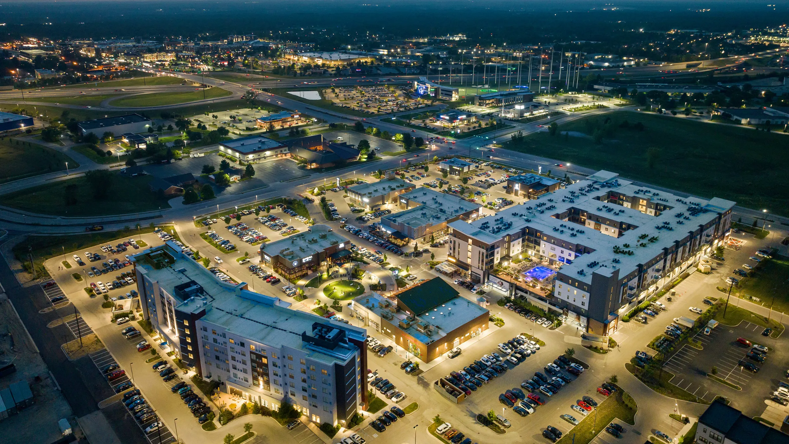 Aerial view of a vibrant, illuminated apartment complex featuring modern architecture, surrounded by ample parking and retail spaces in a bustling neighborhood.