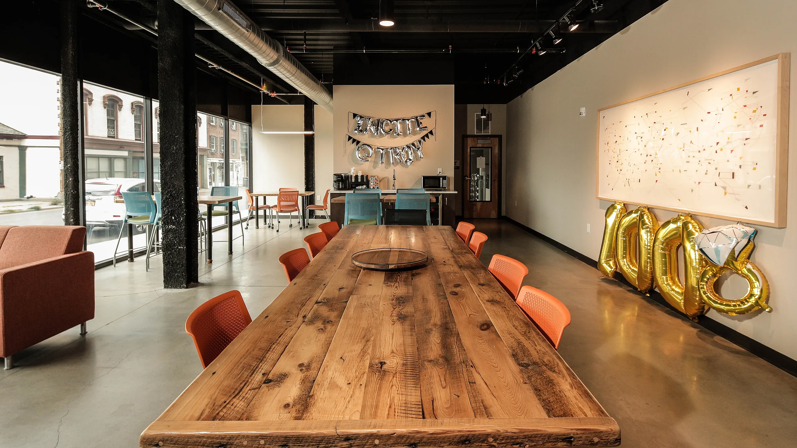 Modern event space featuring a long wooden table with vibrant orange chairs, industrial-style ductwork, and expansive windows offering ample natural light. Perfect for gatherings and celebrations.