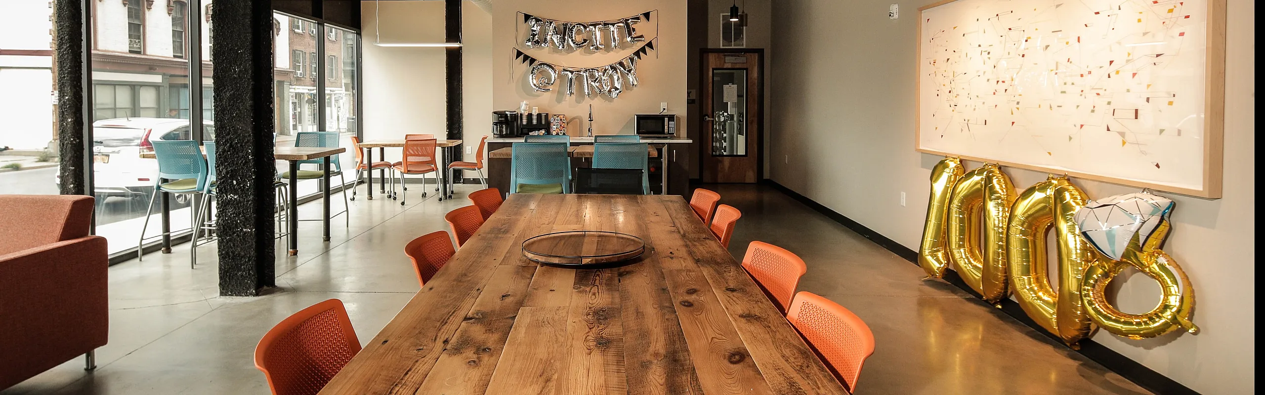 Chic and inviting communal space with a long wooden table, vibrant orange chairs, large windows, and modern decor.