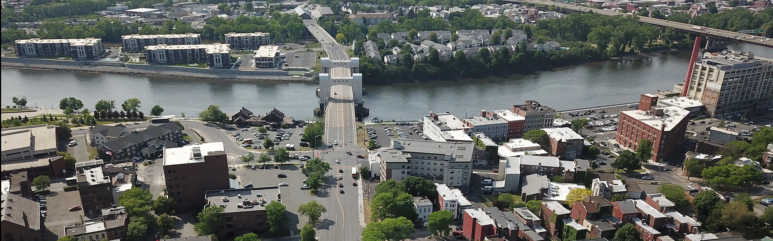 Breathtaking aerial view of a vibrant riverside community with lush greenery, modern buildings, and convenient roadway access.