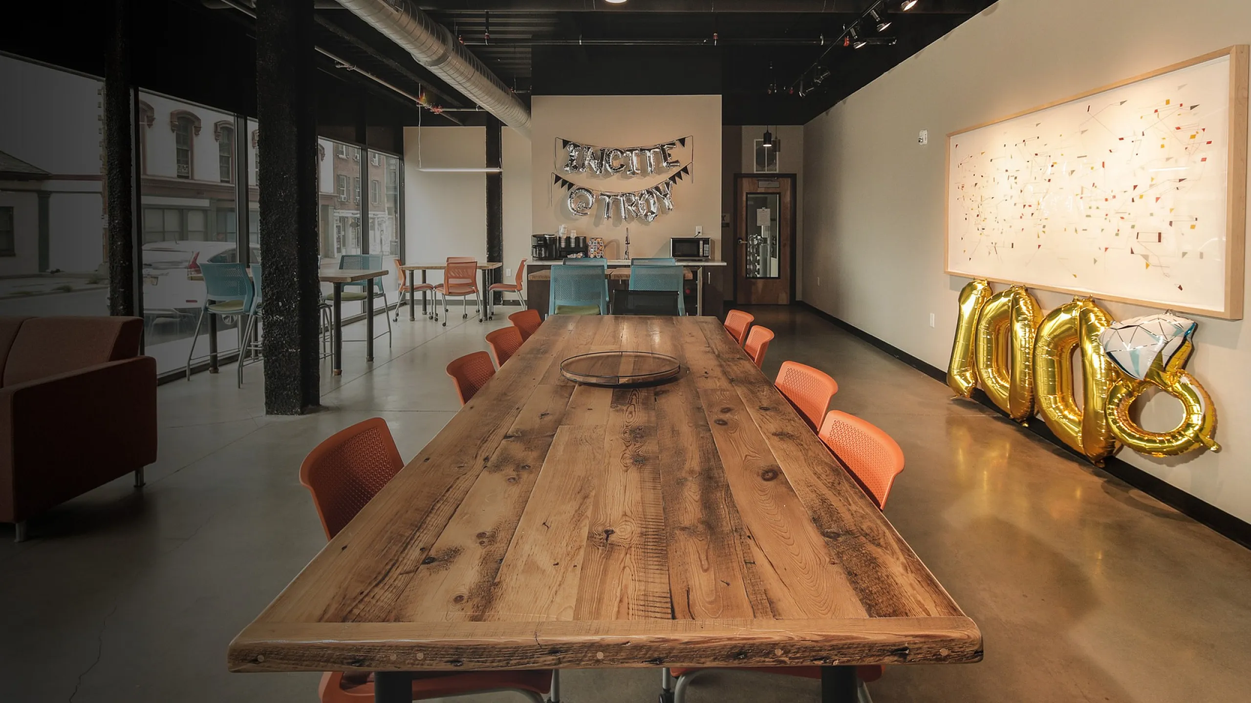 Chic modern workspace with rustic wooden table, vibrant orange chairs, and creative wall decor under industrial lighting.