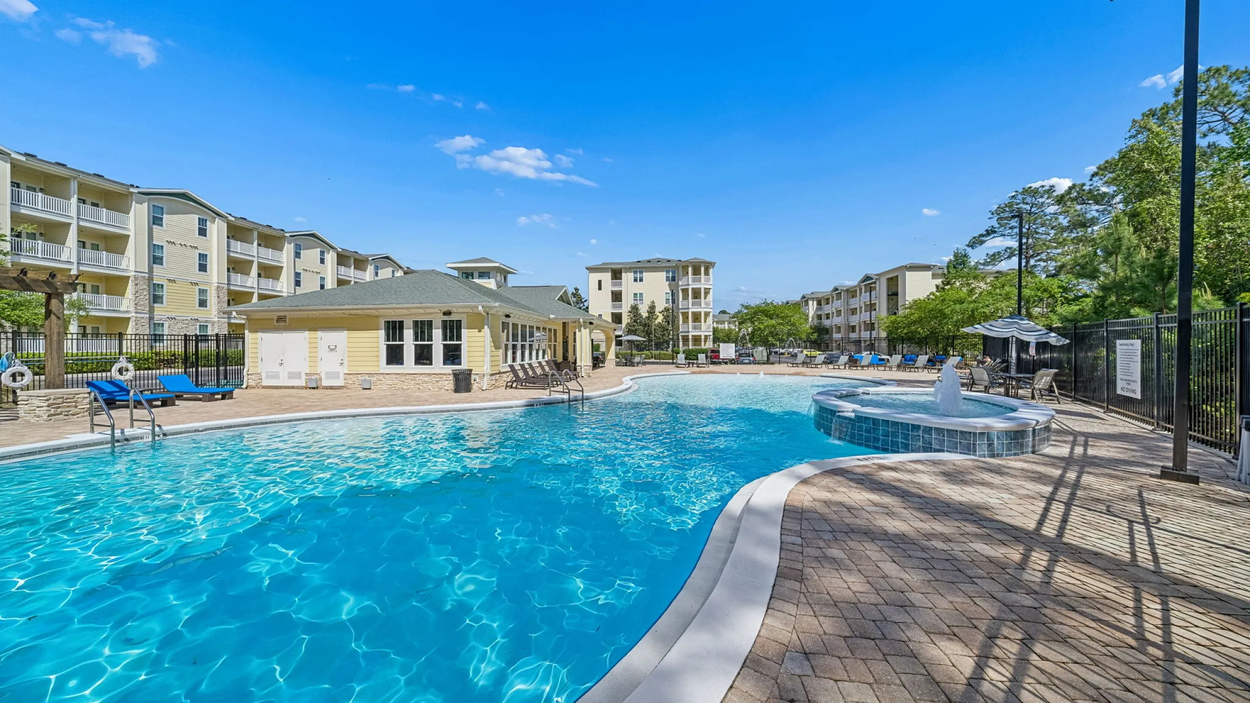 Inviting apartment community featuring a luxurious pool area with lounge chairs, surrounded by modern buildings under a clear blue sky.