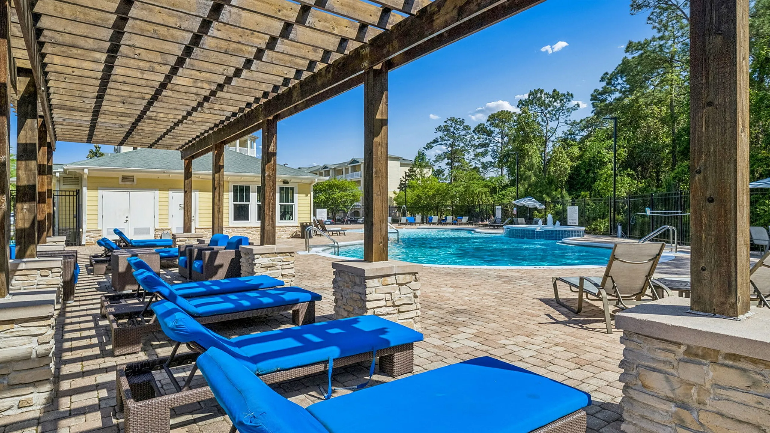 Luxurious poolside area featuring vibrant blue lounge chairs under a rustic pergola, surrounded by lush greenery and modern amenities.