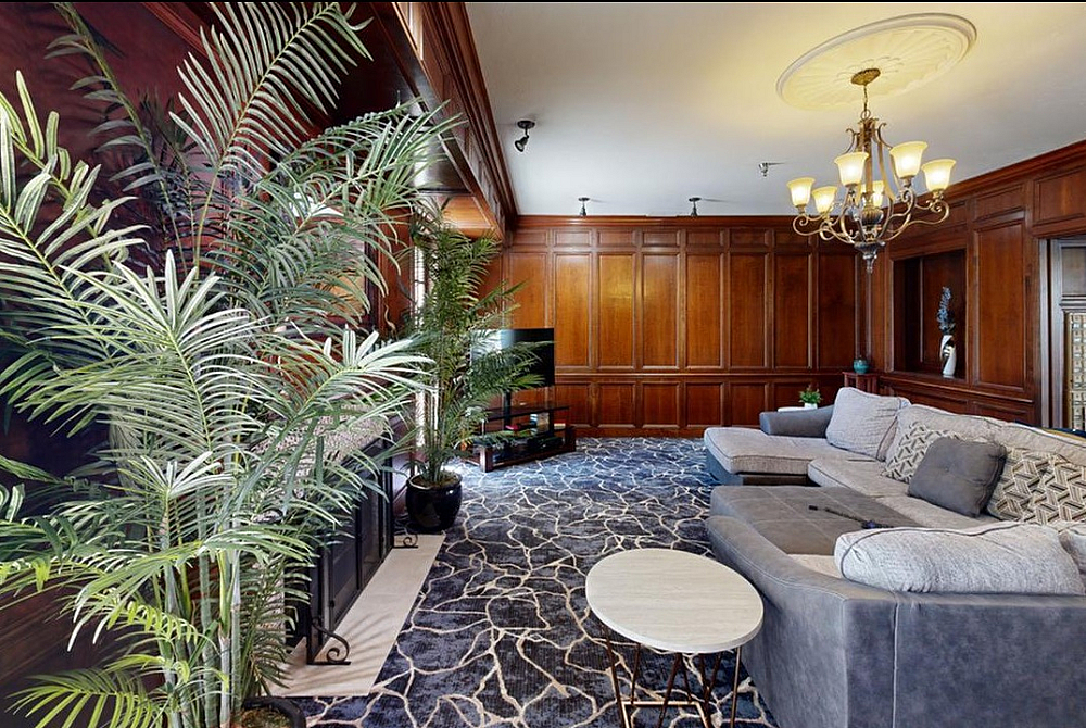 Elegant living room with rich wooden paneling, plush gray sectional, and vibrant green plants, featuring a stylish chandelier and patterned carpet for a luxurious feel.