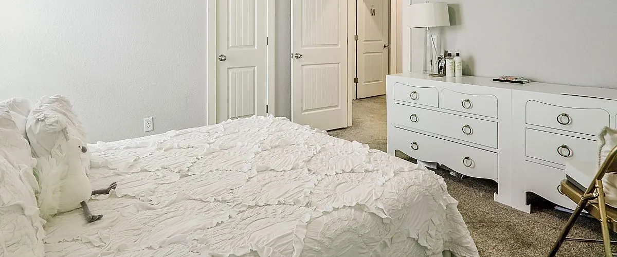 Bright and elegant bedroom with light gray walls, cozy bed with textured white bedding, and a sleek ceiling fan. Features ample natural light from the large window and stylish white furniture.