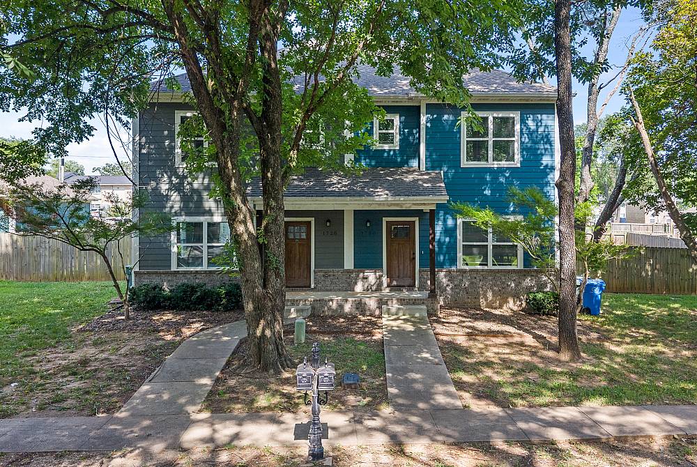 Charming duplex with blue siding, nestled among lush trees, offering inviting front entrances and ample outdoor space.