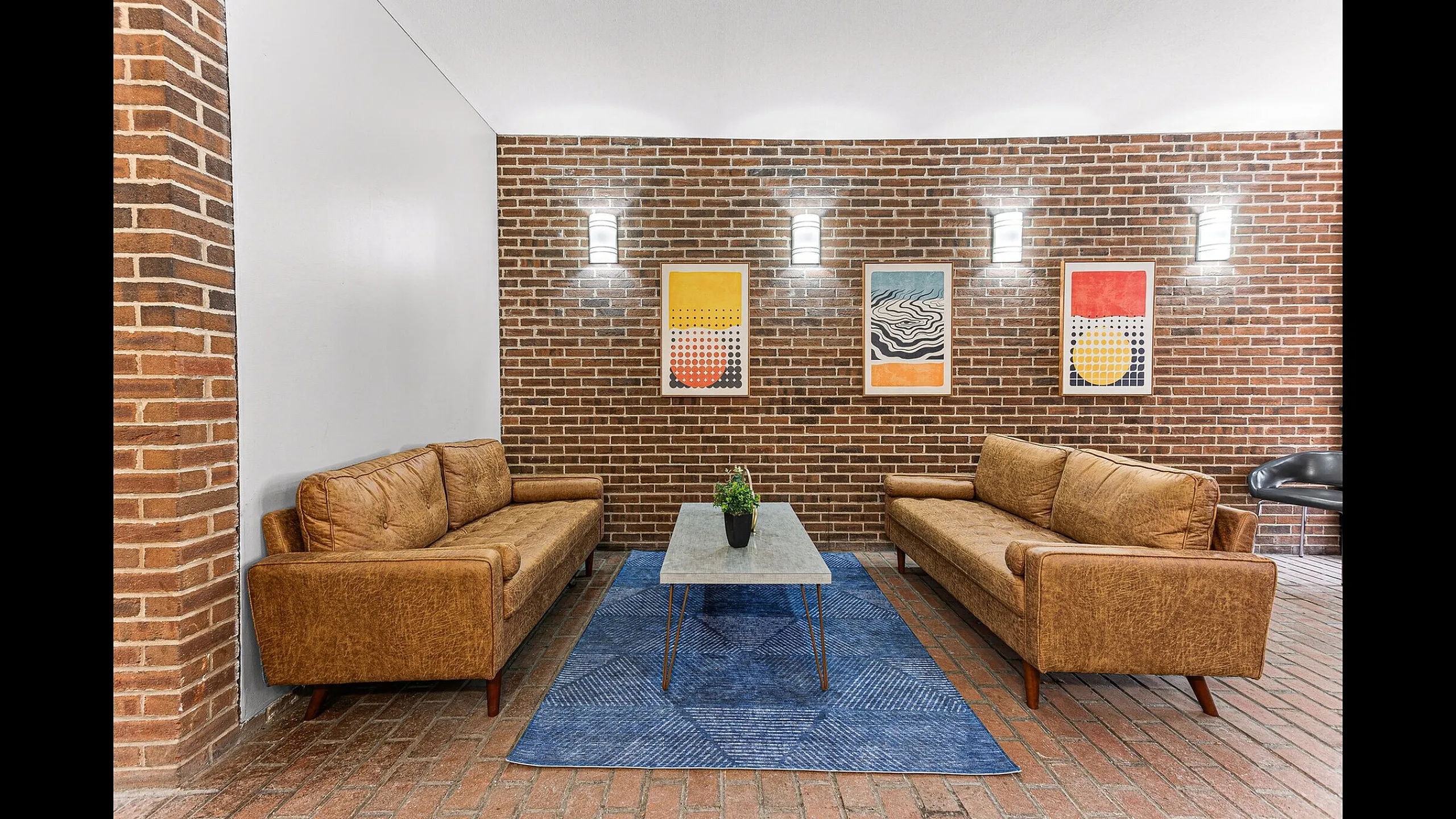Chic brick-walled living area featuring cozy leather sofas, vibrant abstract art, and a modern coffee table on a stylish blue rug.