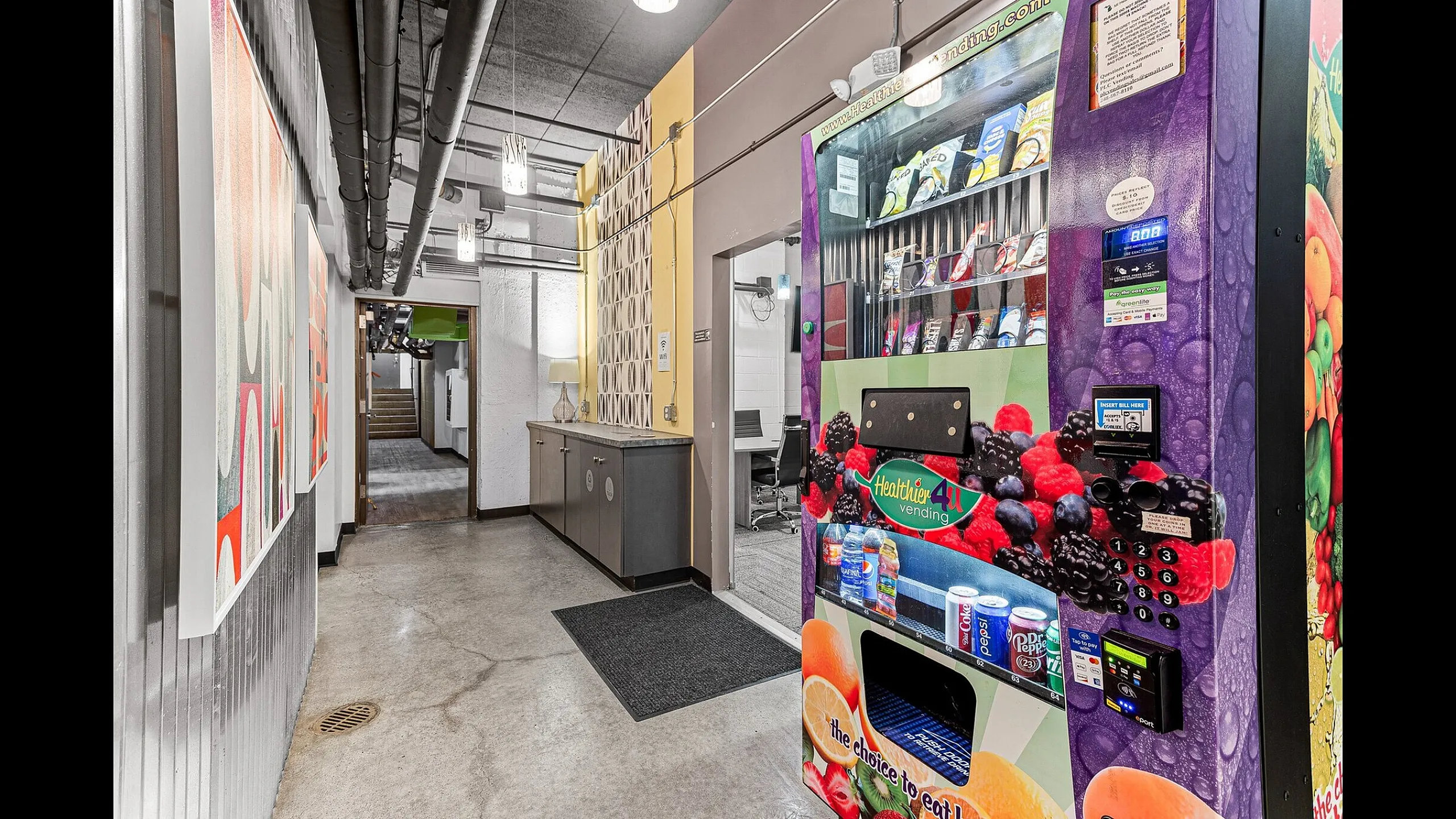 Modern hallway with vibrant art, sleek vending machine, and industrial-chic design leading to office space.