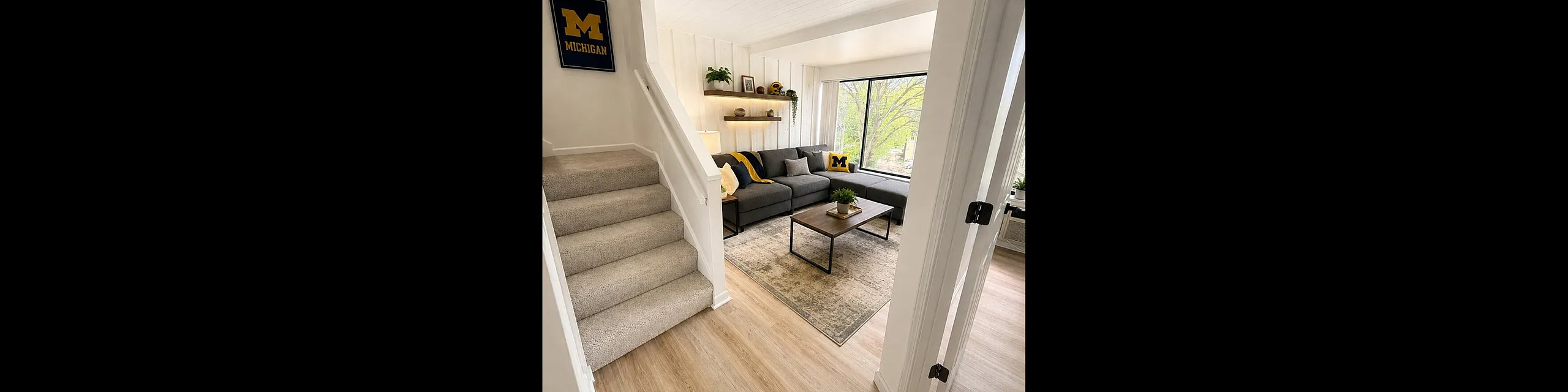 Inviting living room with cozy gray sectional, stylish wooden coffee table, and large window offering abundant natural light, accented by Michigan-themed decor and plush carpeted stairs.