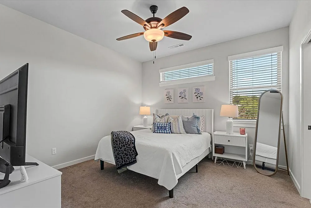 Bright and modern bedroom with elegant decor, featuring a cozy bed with plush pillows, ceiling fan, stylish bedside lamps, and a sleek full-length mirror, bathed in natural light.