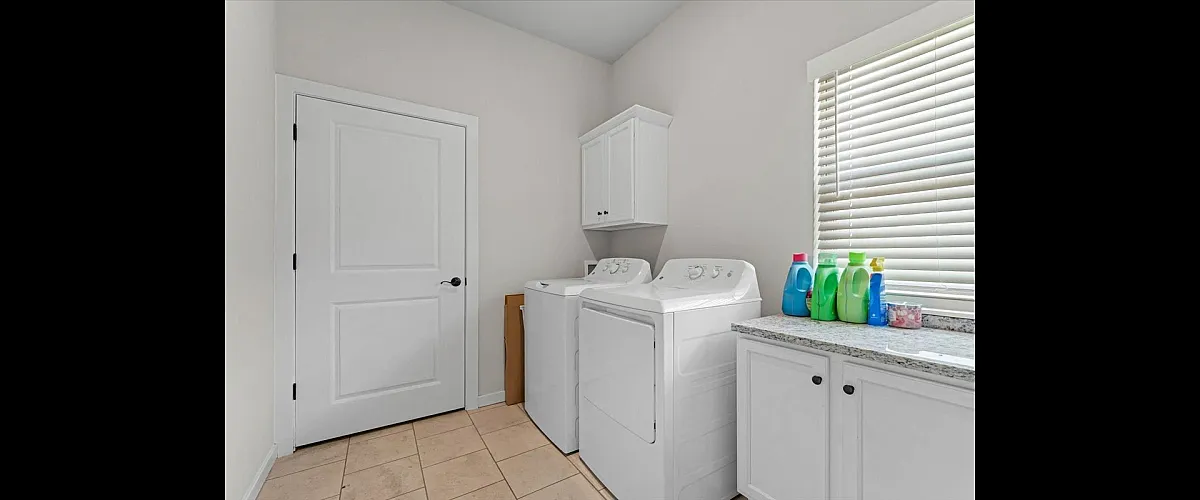 Bright and functional laundry room with modern washer and dryer, ample cabinet storage, and a sunlit window for natural light.