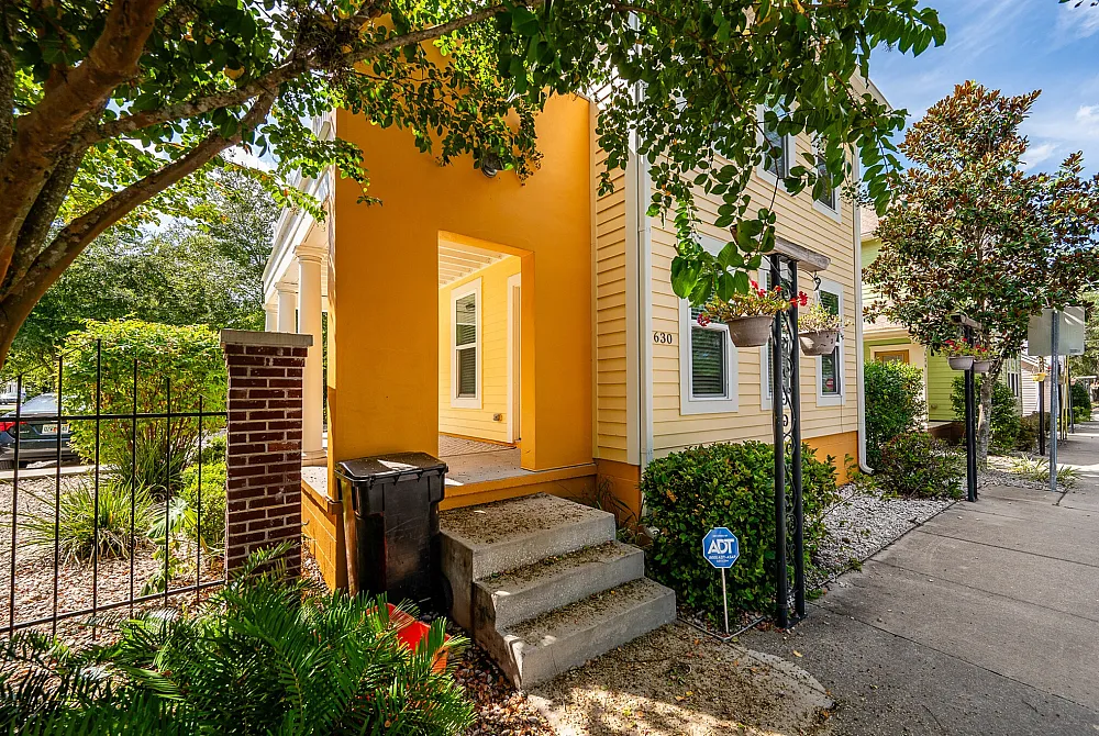 Charming yellow house exterior with inviting entryway, surrounded by lush greenery and vibrant hanging plants.
