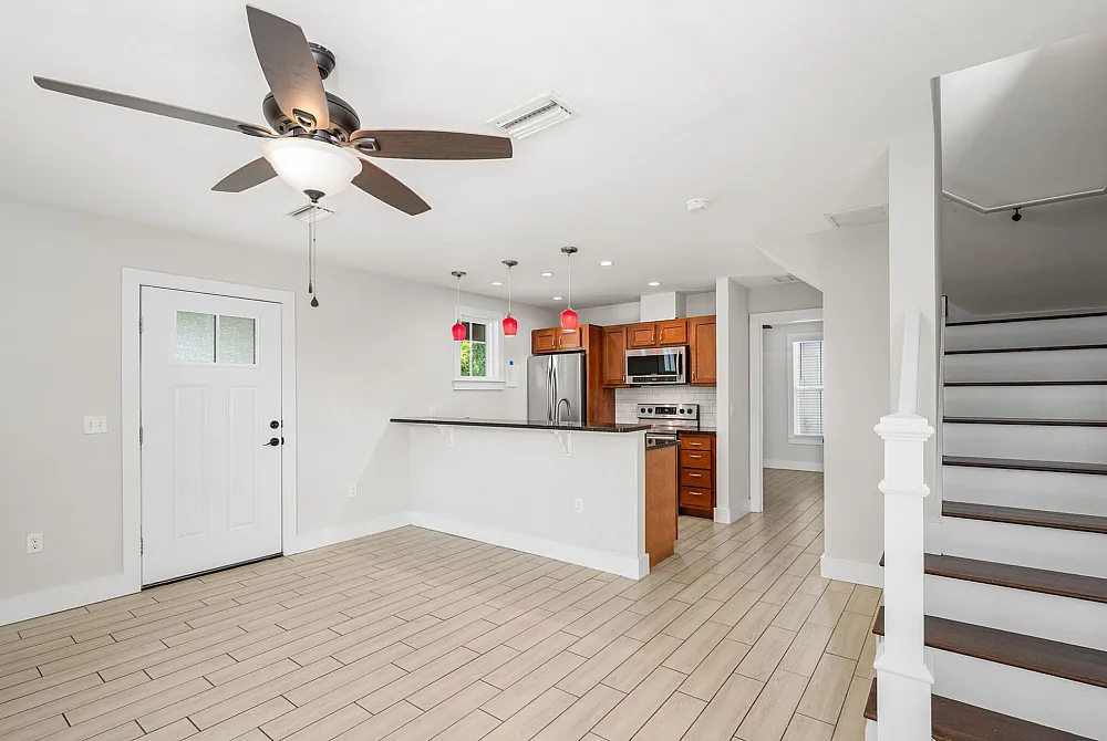 Bright and modern open-concept kitchen with sleek stainless steel appliances, stylish wooden cabinetry, and elegant pendant lighting, complemented by a spacious living area and contemporary ceiling fan.