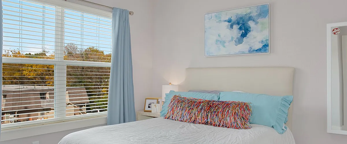 Bright and elegant bedroom with light gray walls, cozy bed with textured white bedding, and a sleek ceiling fan. Features ample natural light from the large window and stylish white furniture.