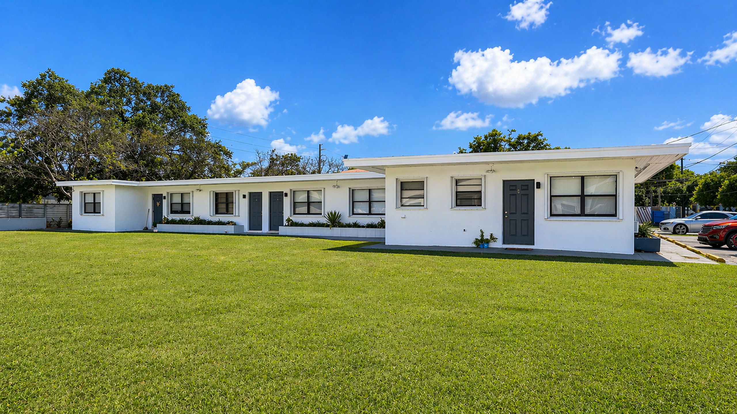 Charming single-story white apartment complex with modern doors, surrounded by lush green lawn and beautiful blue skies.