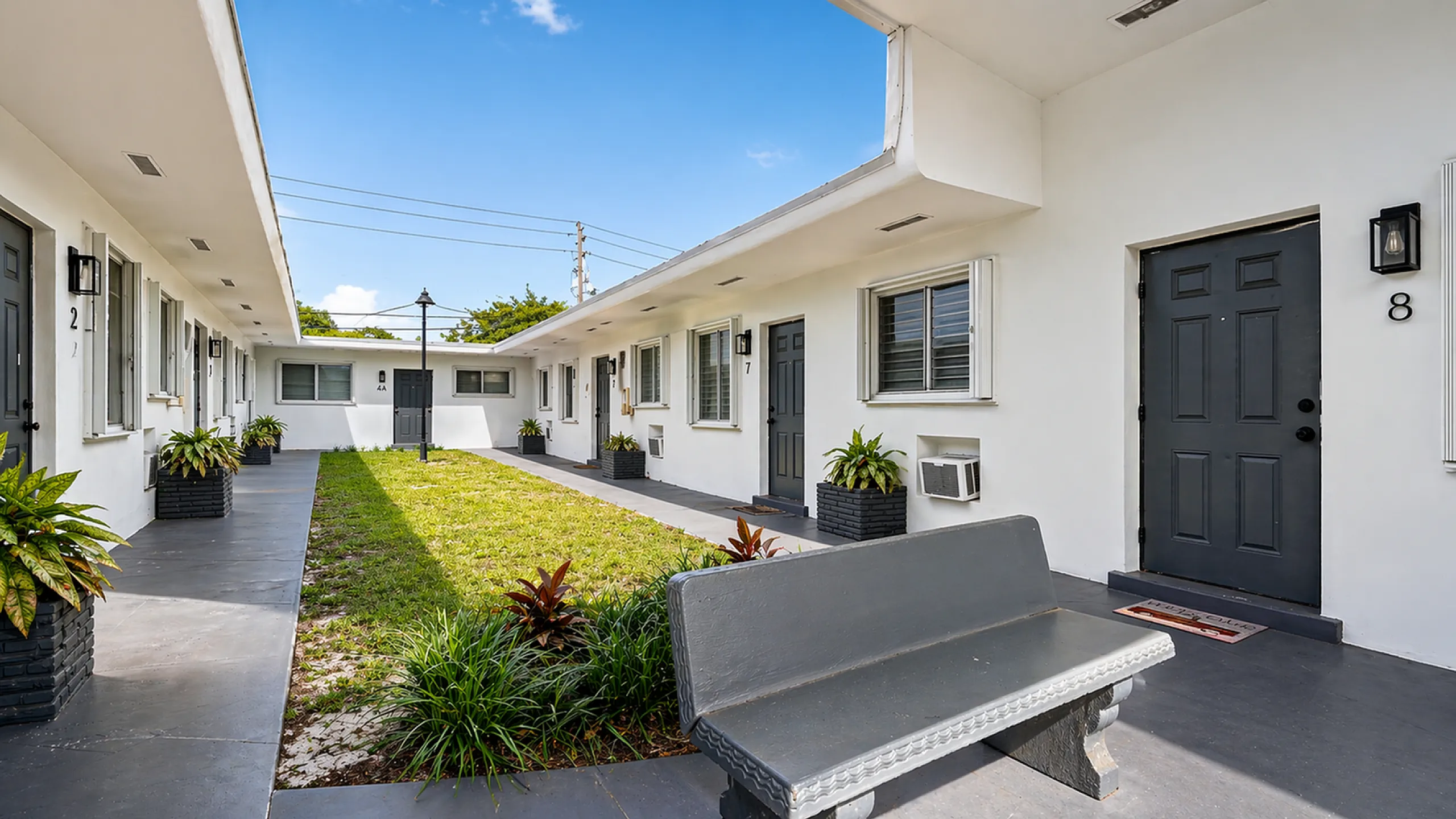 Charming white apartment complex with modern black doors, featuring a lush green courtyard and elegant potted plants, perfect for comfortable living.