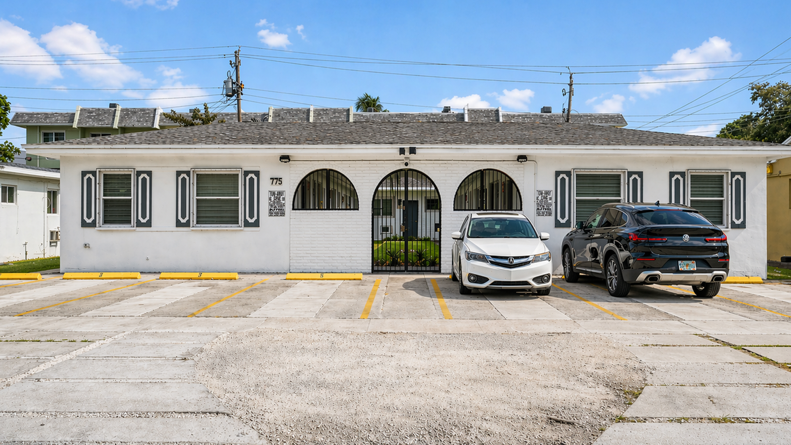 Charming single-story apartment complex with classic white facade and decorative arched windows, featuring ample parking space.