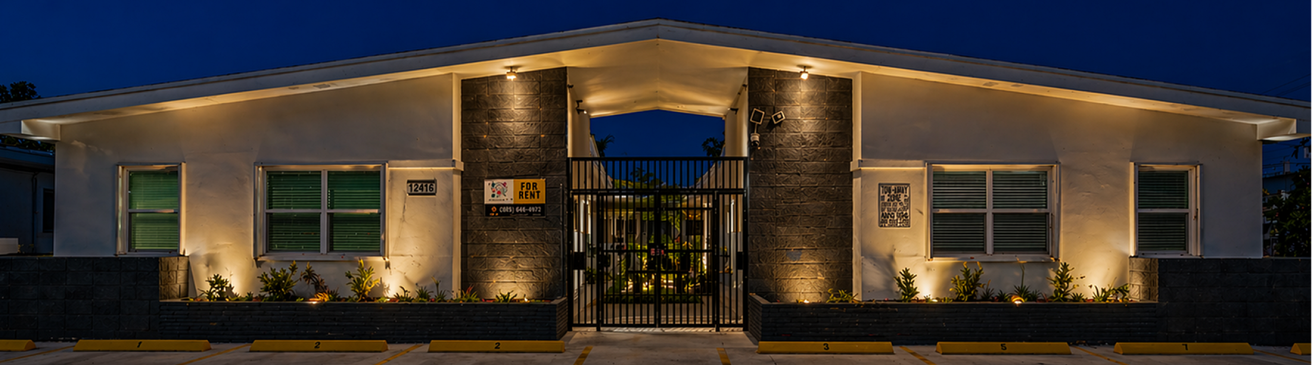Elegant modern apartment exterior with warm evening lighting, lush landscaping, and inviting entrance for rent.
