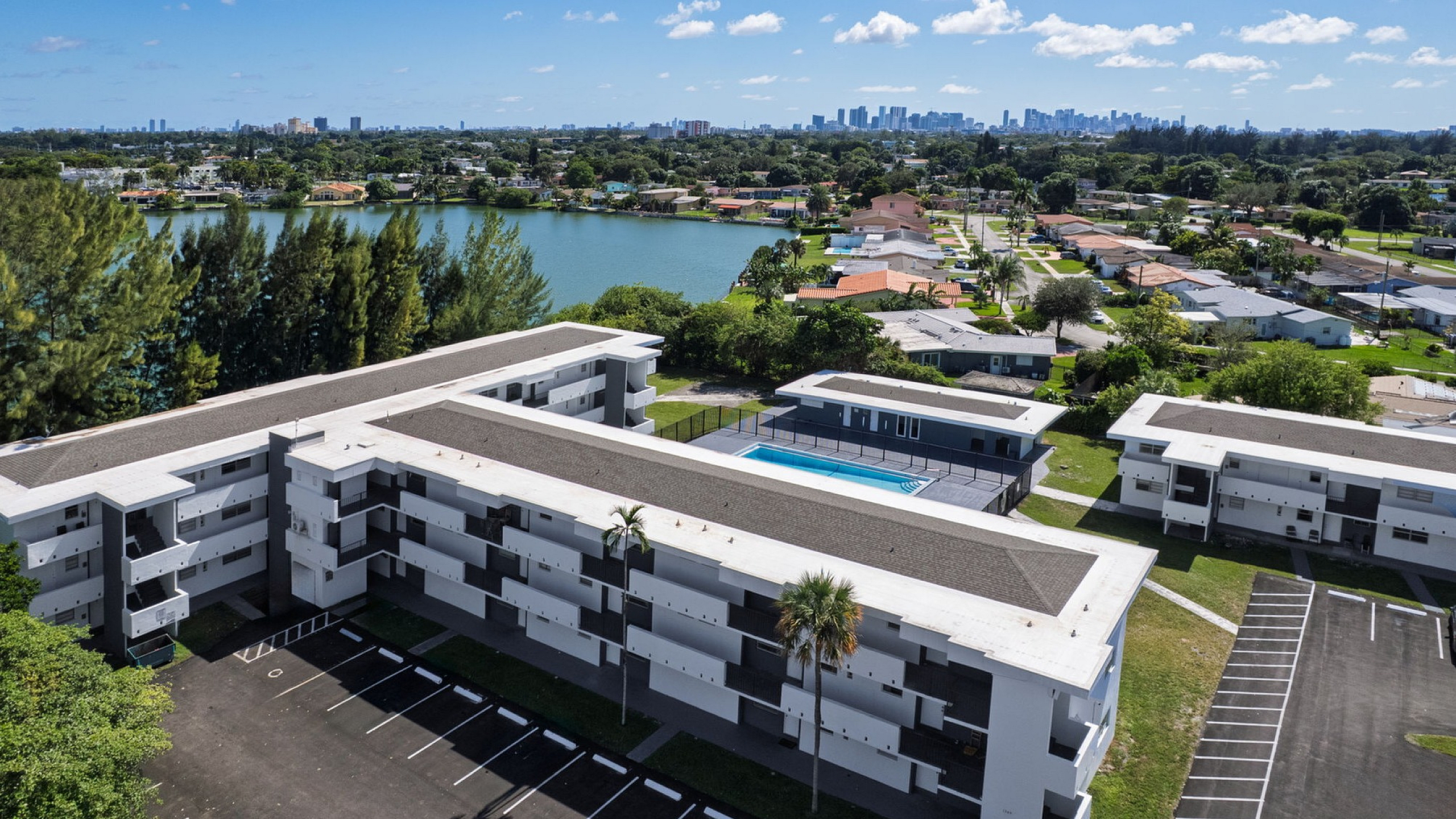 Scenic aerial view of a modern apartment complex with a central pool, surrounded by lush greenery and a serene lake, offering tranquil cityscape views.